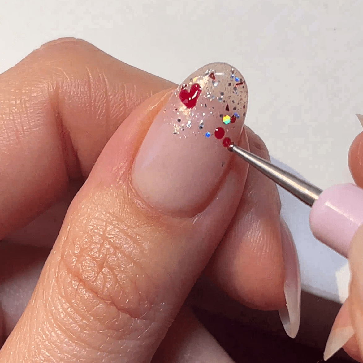 A hand paints red dots on a nail with glitter, using a fine brush.
