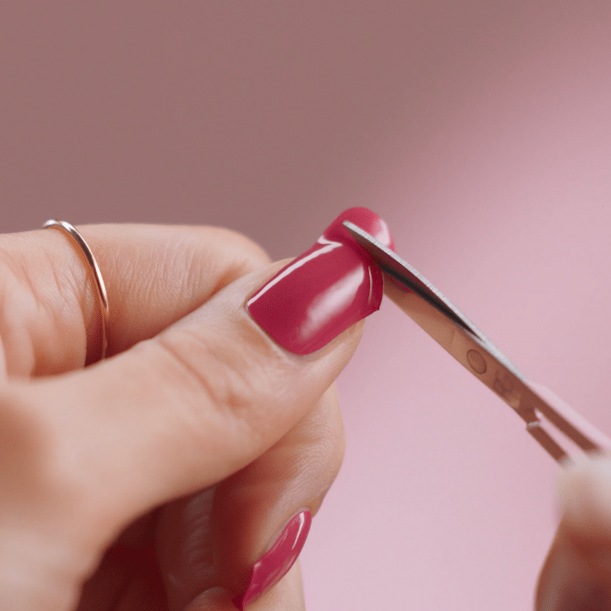 Close-up of a hand holding a pink painted nail using nail scissors against a pink background.