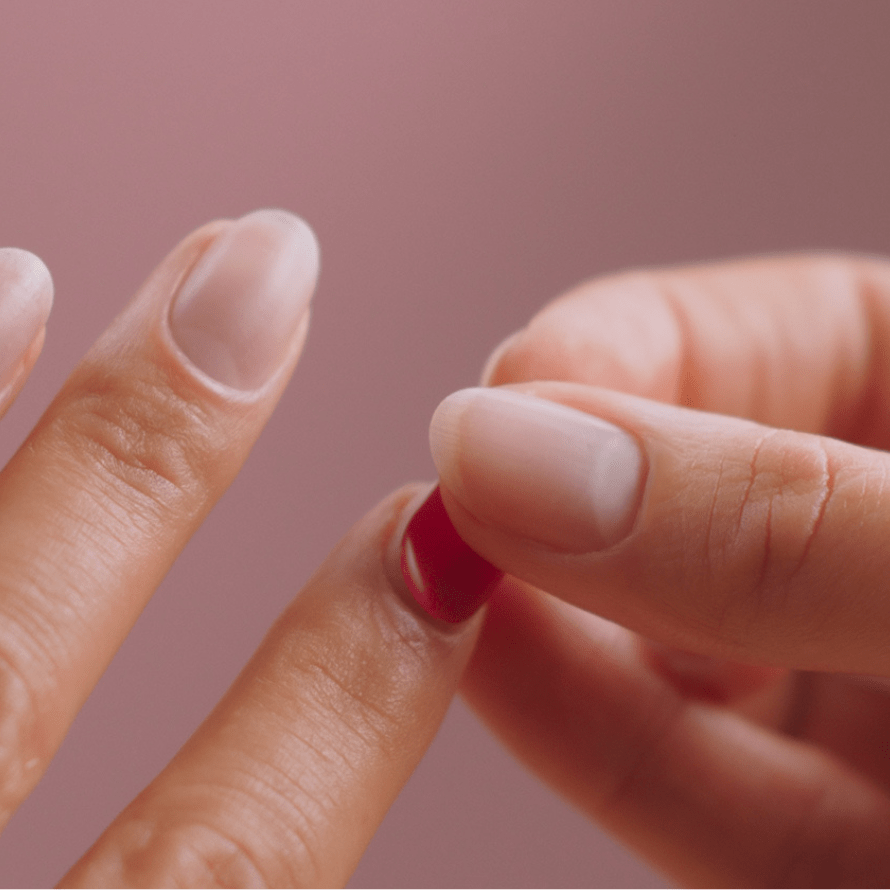 Close-up of hands placing a red tacknail on a fingernail, against a pink background.