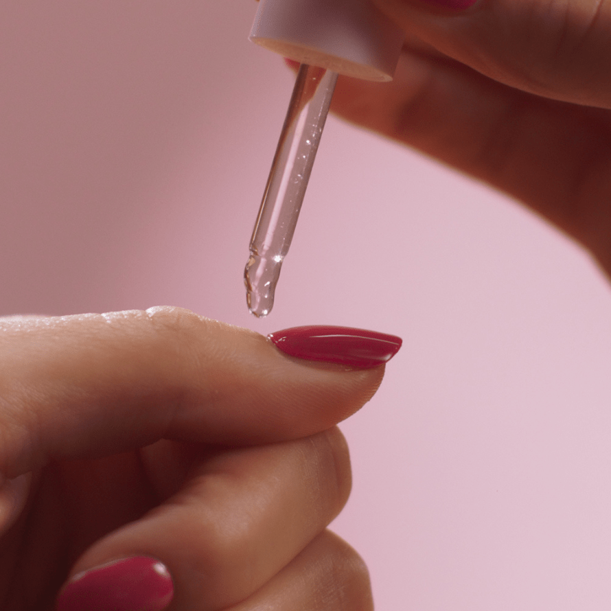 A hand with pink painted nails drips a liquid on the nail with a pipette against a pink background.