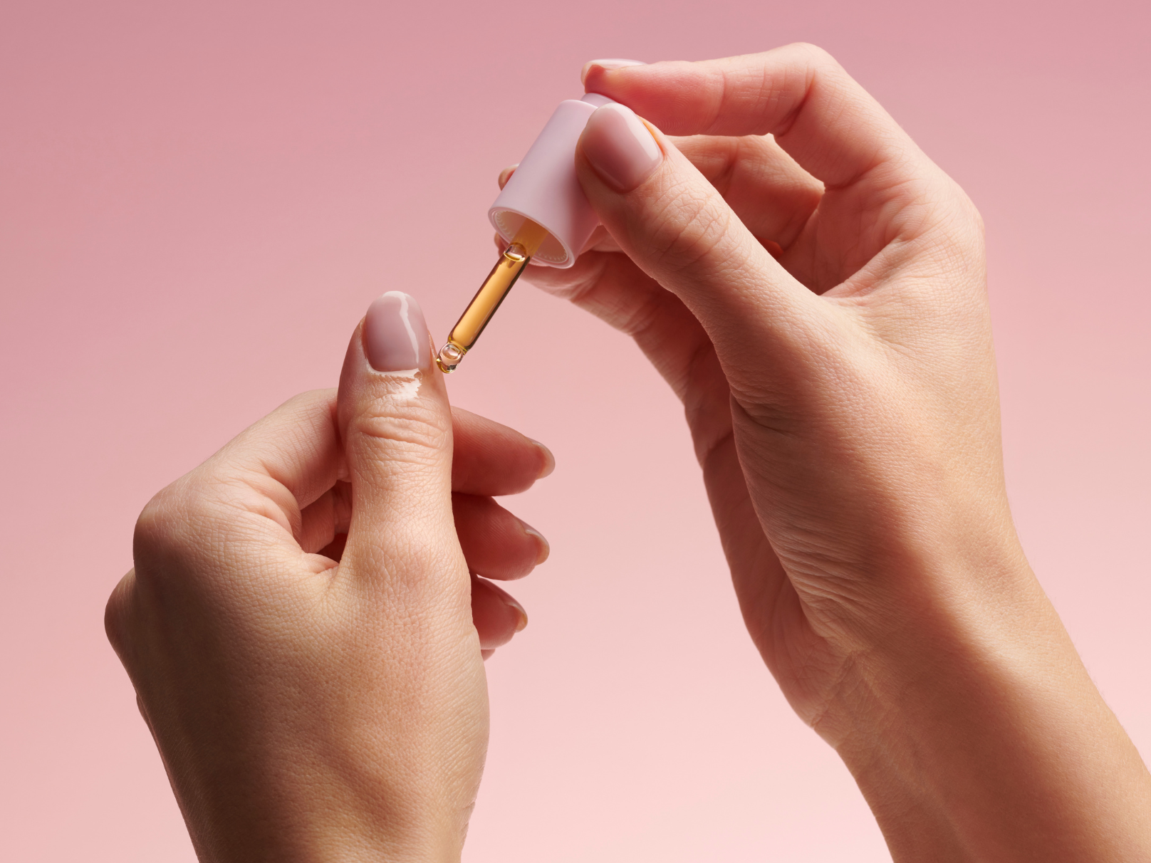 Two hands with well-groomed nails use a dropper with an oily liquid against a pink background.