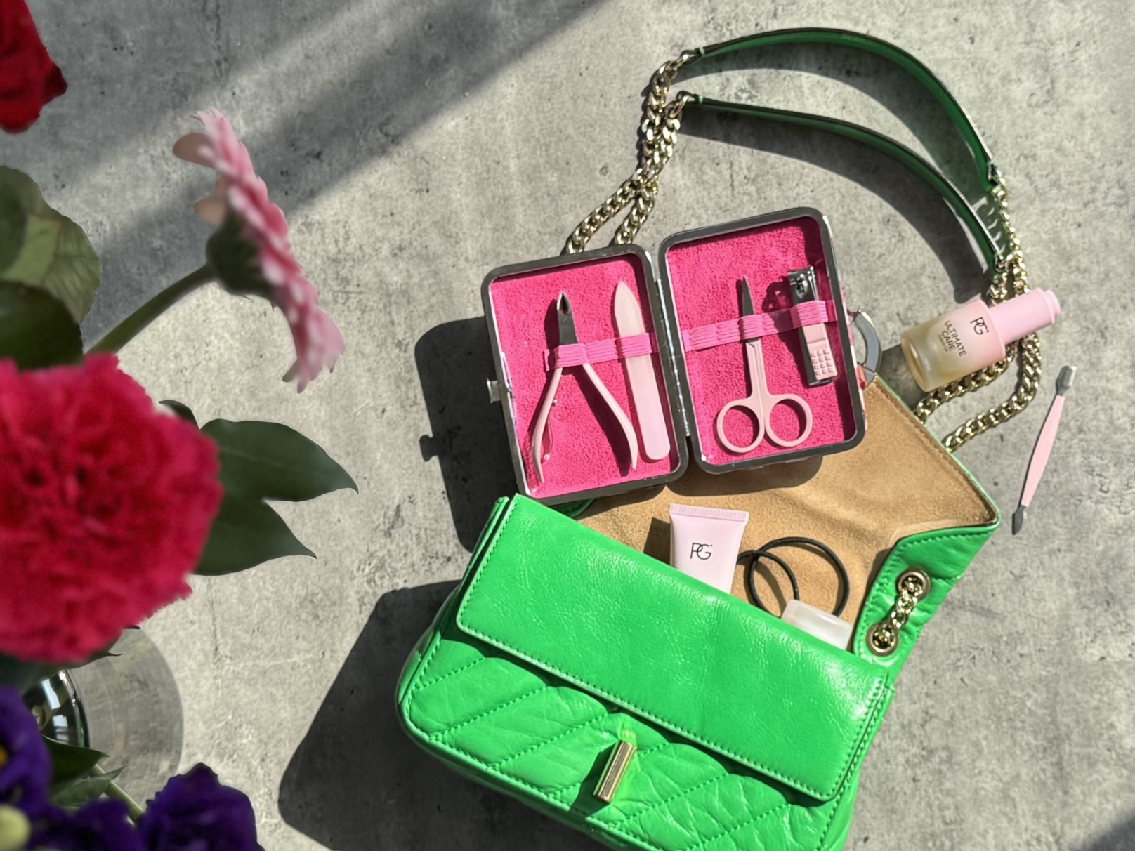 Open green handbag with manicure kit, skincare products and flowers on a concrete surface.