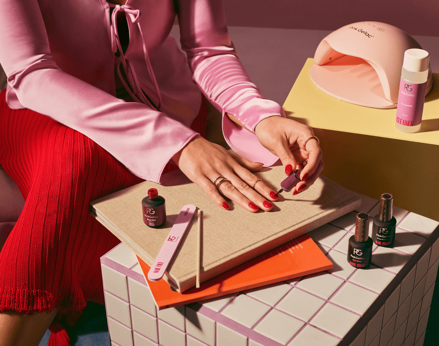 Person with pink blouse doing manicure with red nail polish surrounded by nail care products on a tile surface.