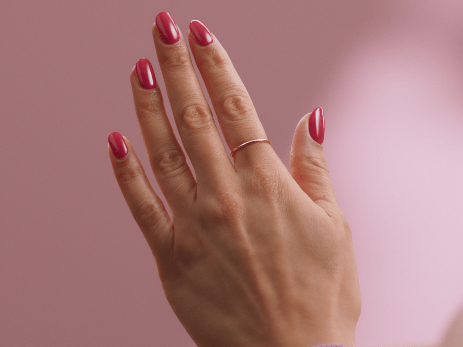A hand with bright pink painted nails and a thin gold ring against a pink background.
