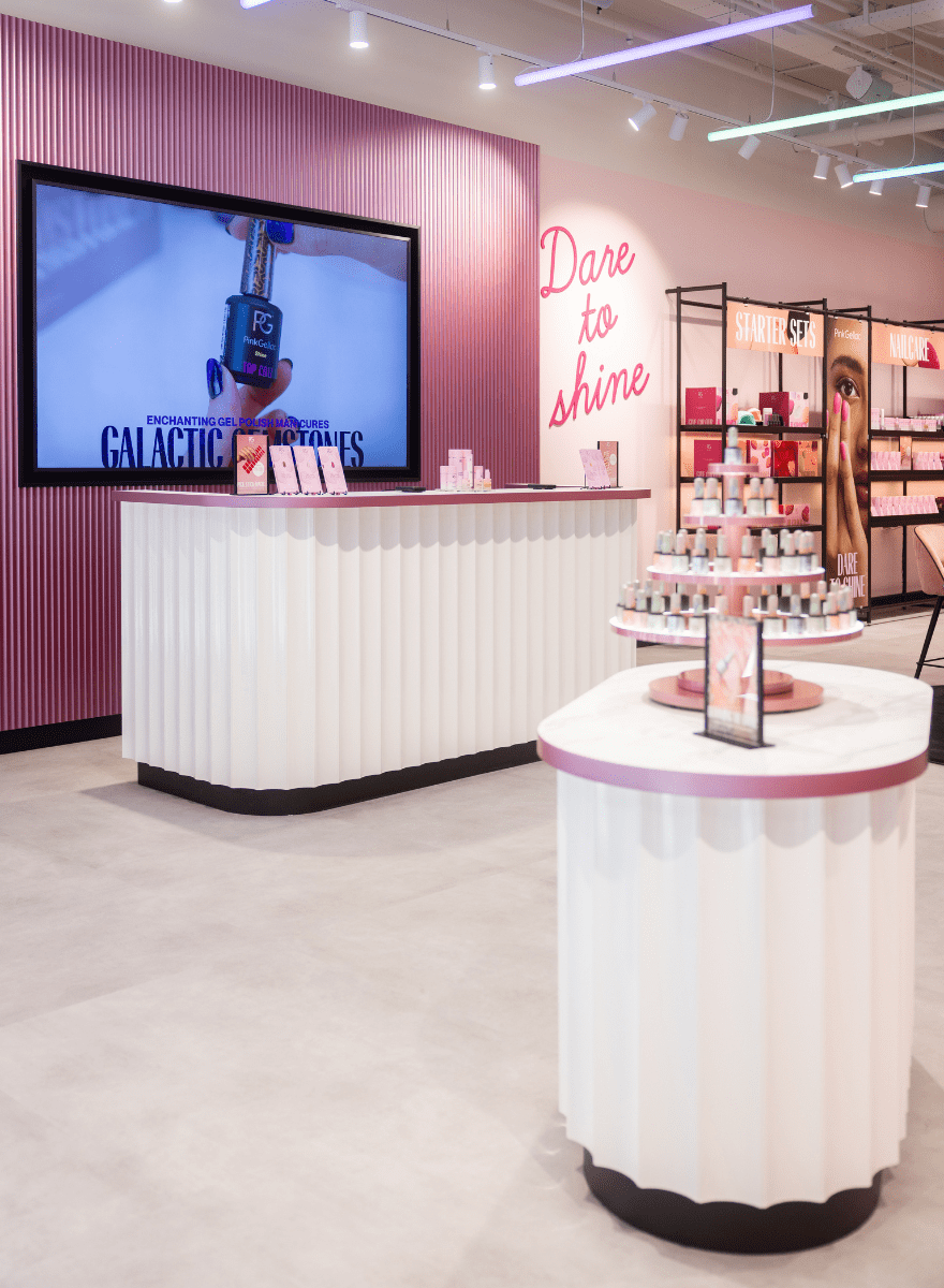 A modern store with a white and pink interior, a large screen on the wall, and displays with nail polish products. The text "Dare to shine" is written on a pink wall.