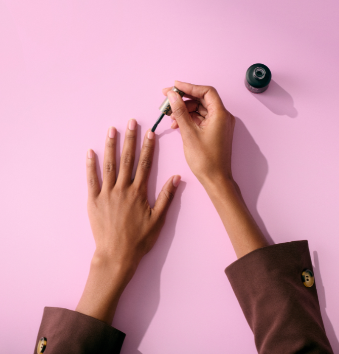 Two hands in brown sleeves paint nails with beige nail polish on a pink background.