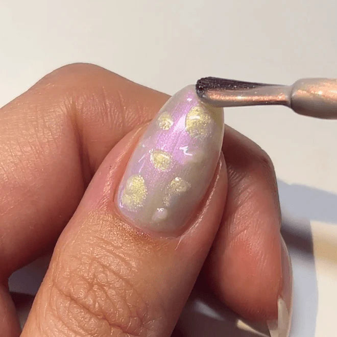 A hand applying a metallic colored nail polish with golden dots to a nail with a brush.