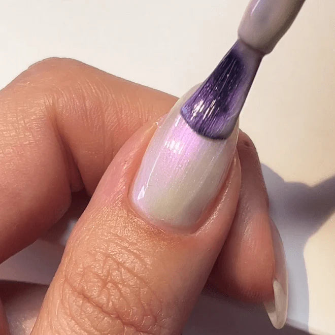 Person applies purple nail polish to a white nail with a brush.