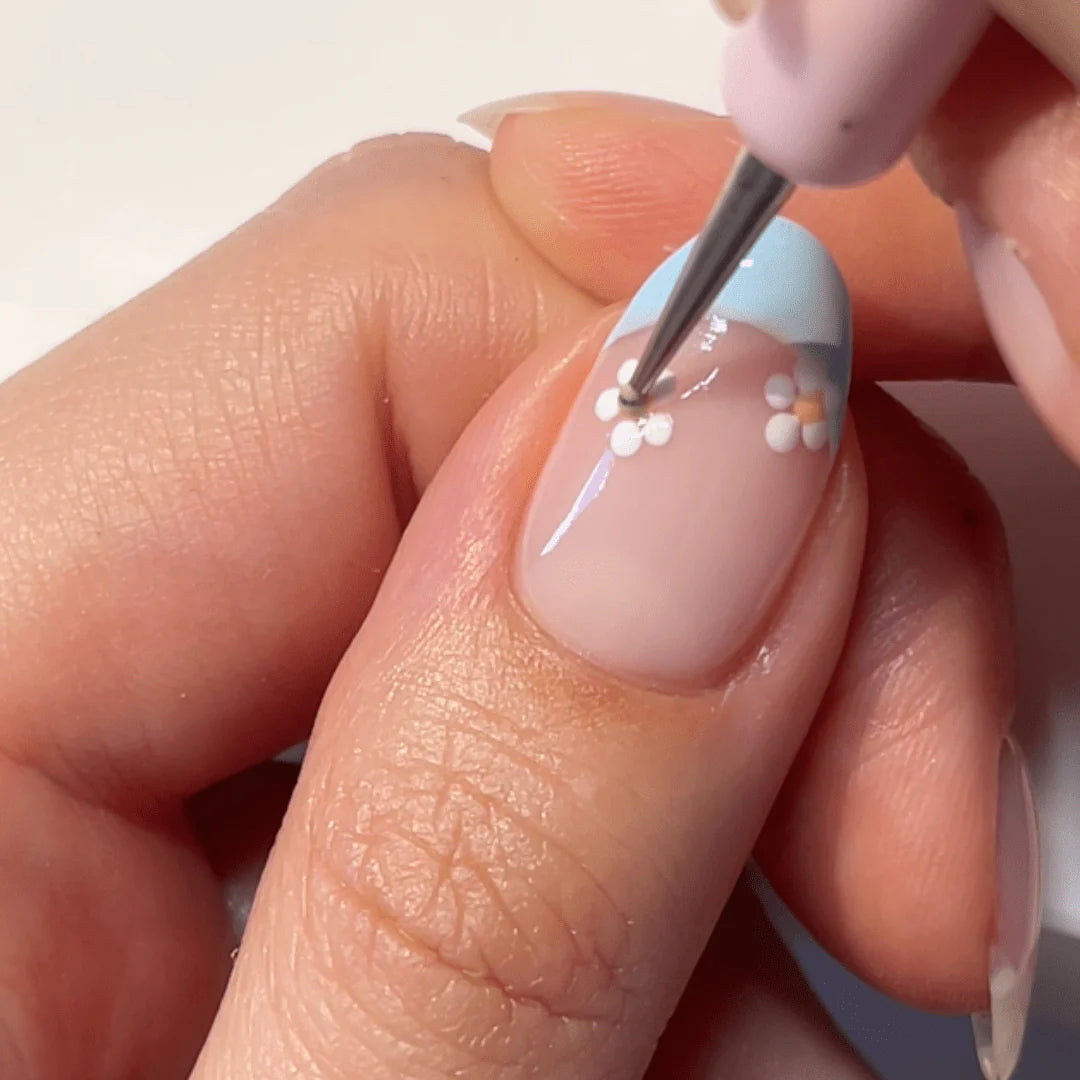 A close-up of a hand painting a light blue nail with small white flowers with a fine brush.
