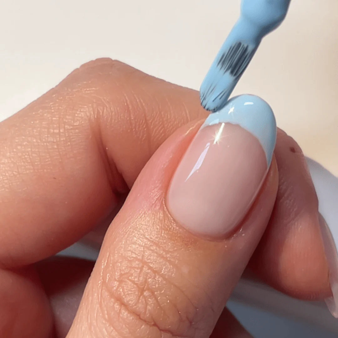 One hand holds a finger while a brush applies light blue nail polish to the nail.