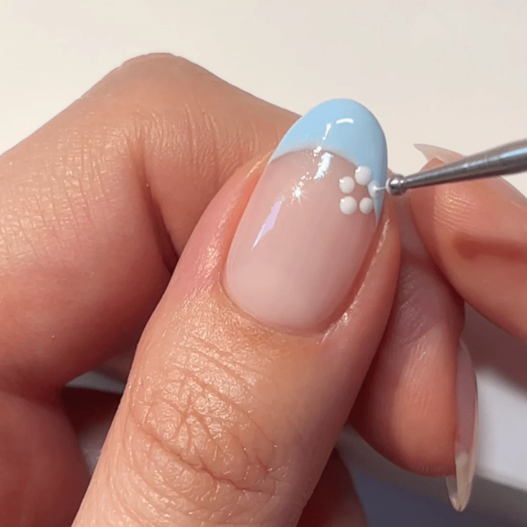 A hand showing a nail with a light blue French manicure and a small white flower decoration on it.