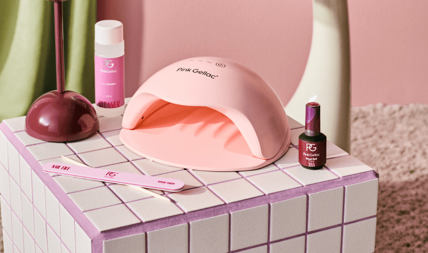 A pink gel polish LED lamp with accessories such as nail polish bottle, cleaner and file, displayed on a tiled table with a pink and green background.