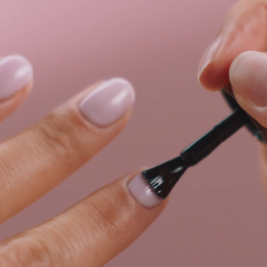 A hand applies light purple nail polish to nails against a pink background.