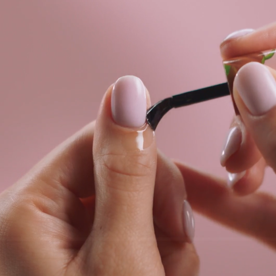 Close-up of a hand applying translucent nail polish to a nail against a pink background.