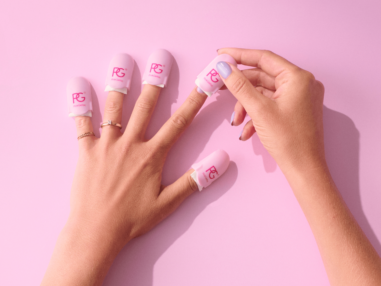 A five-finger hand on which pink nail clips are placed for nail polish removal, against a pink background.