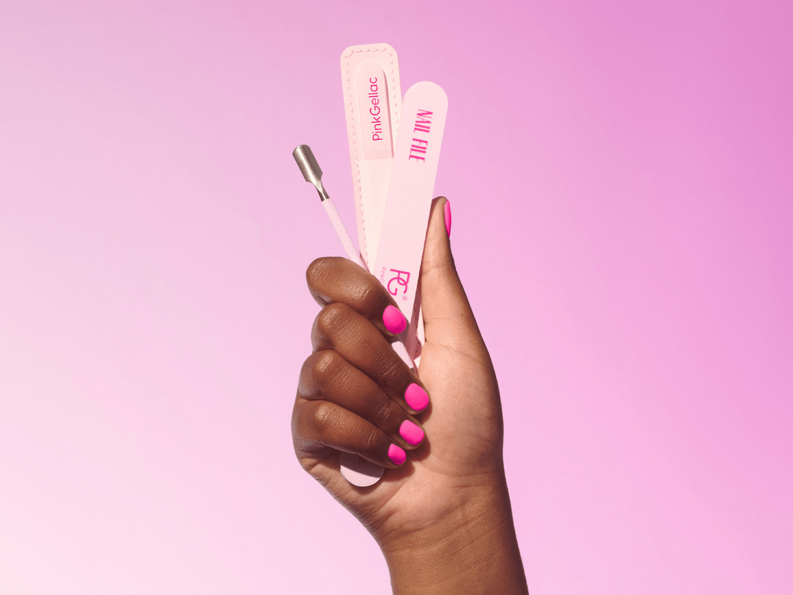 A hand with pink painted nails holds a nail file, a cuticle pusher and another manicure tool against a pink background.