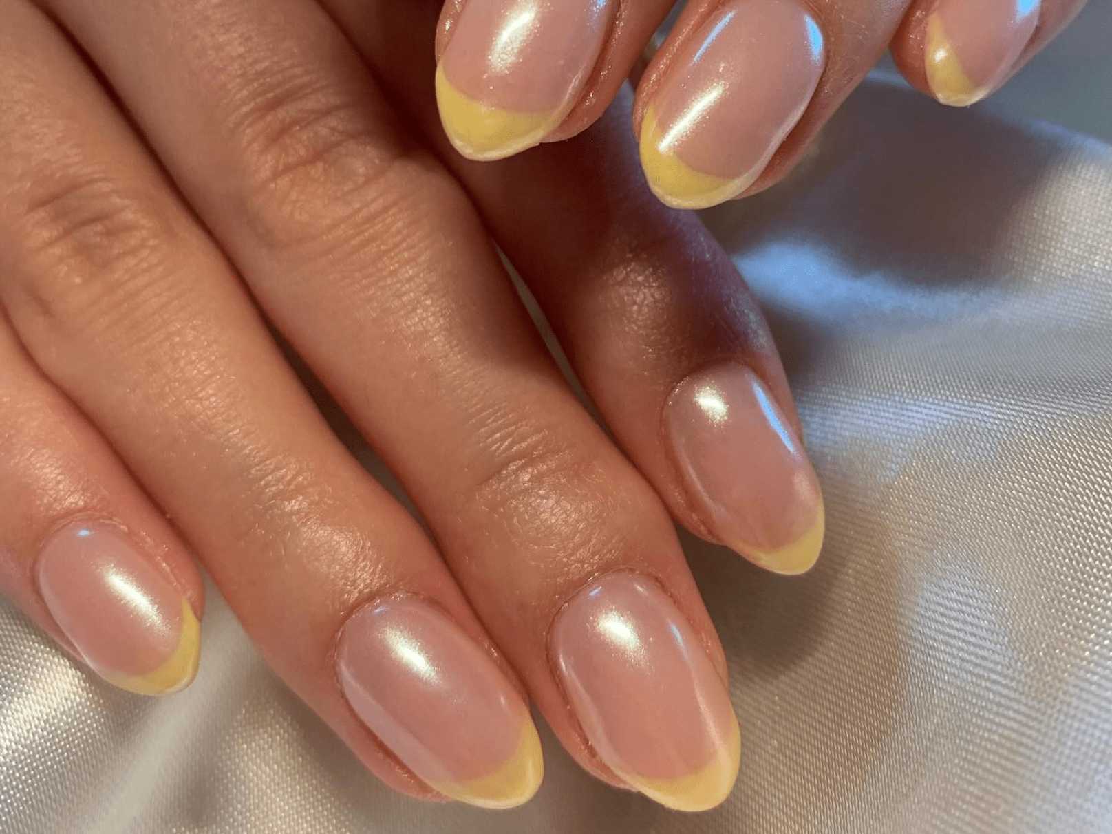 Close-up of hands with shiny nails, polished in pink with a yellow tip, on a satin background.