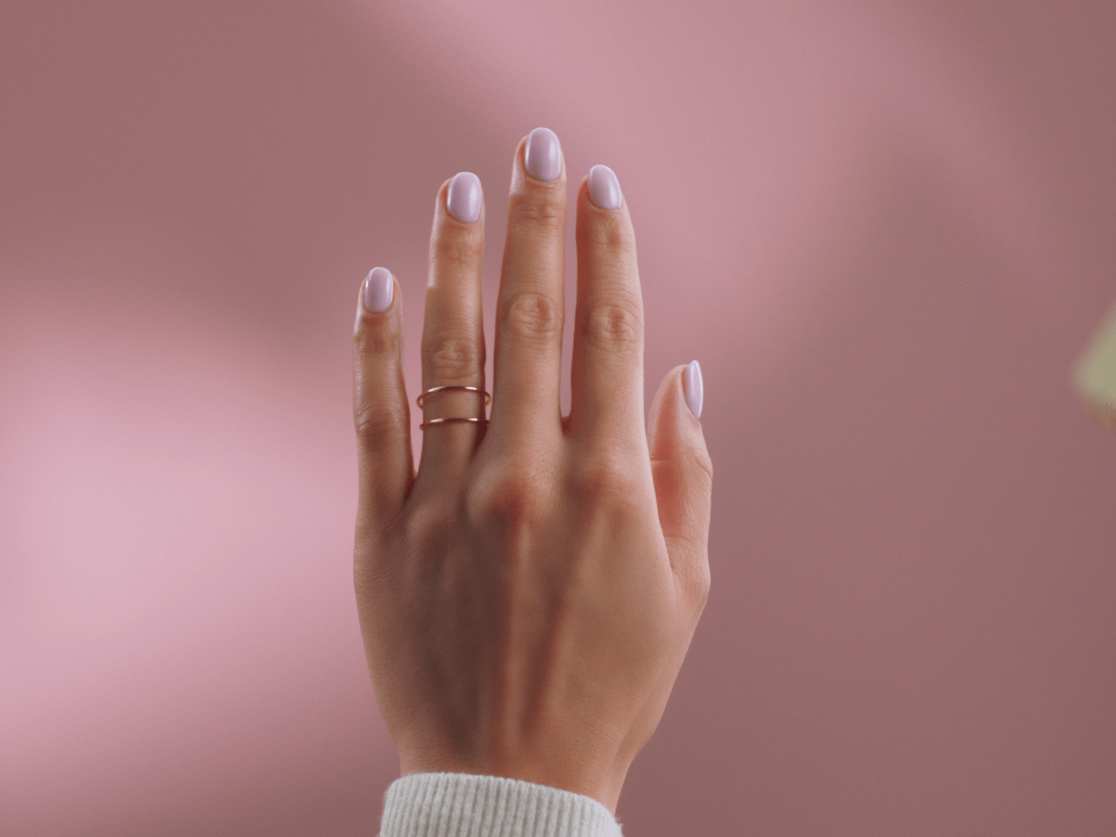 Close-up of a hand with lilac painted nails and a gold ring against a pink background.