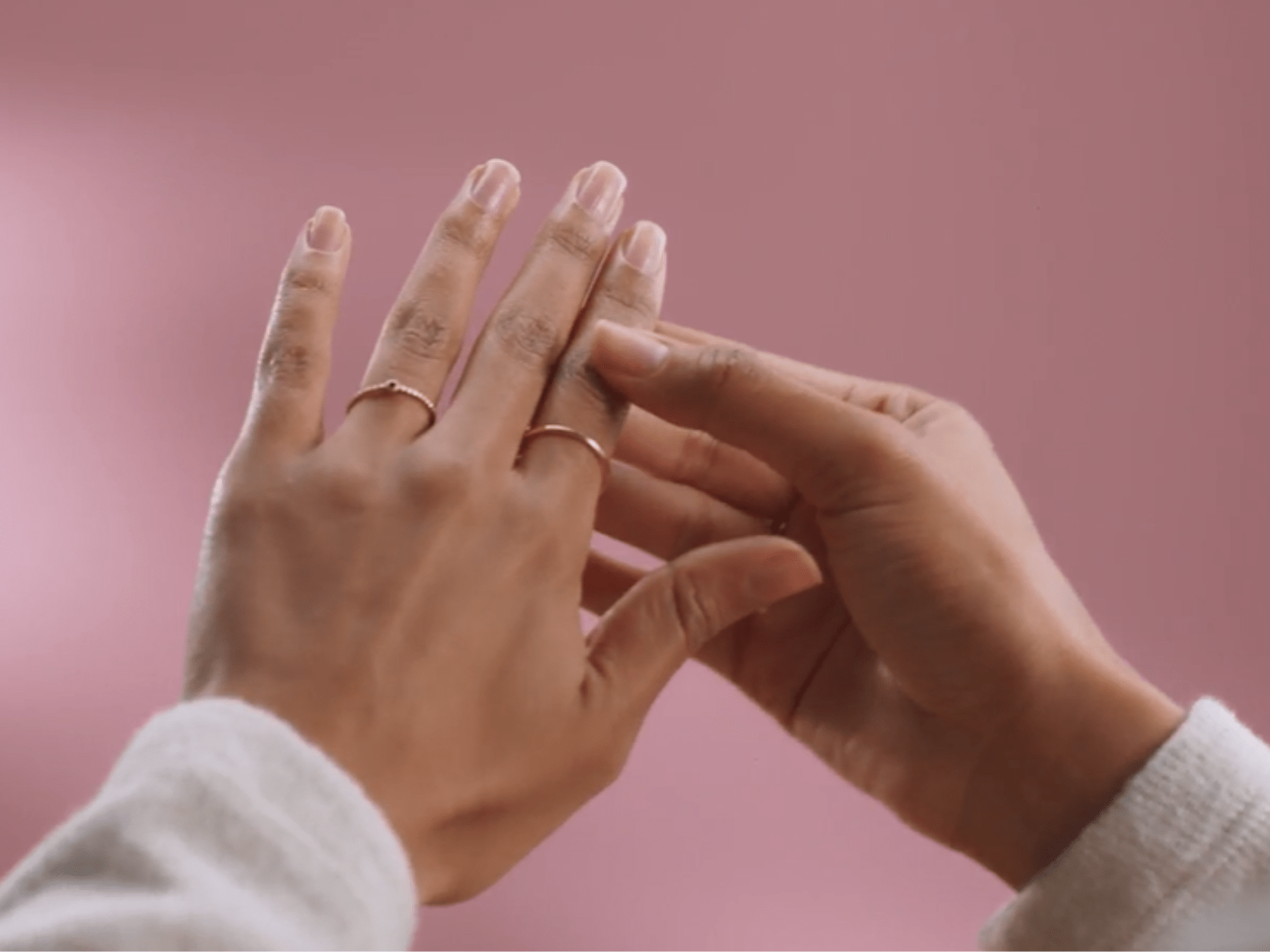 Two hands with pink nail polish and thin gold rings on a pink background.