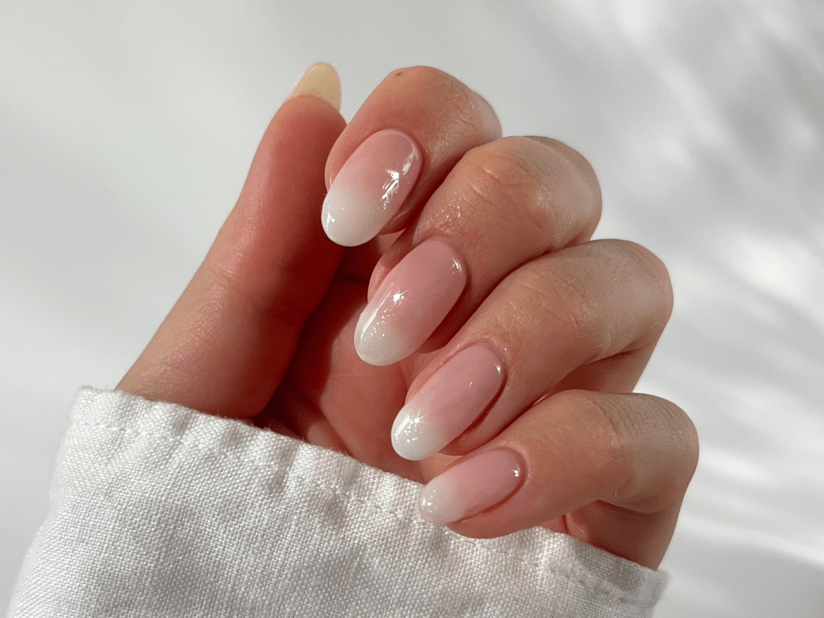 Hand with long, almond-shaped nails, colored in a soft ombre from pink to white, with a glossy finish.