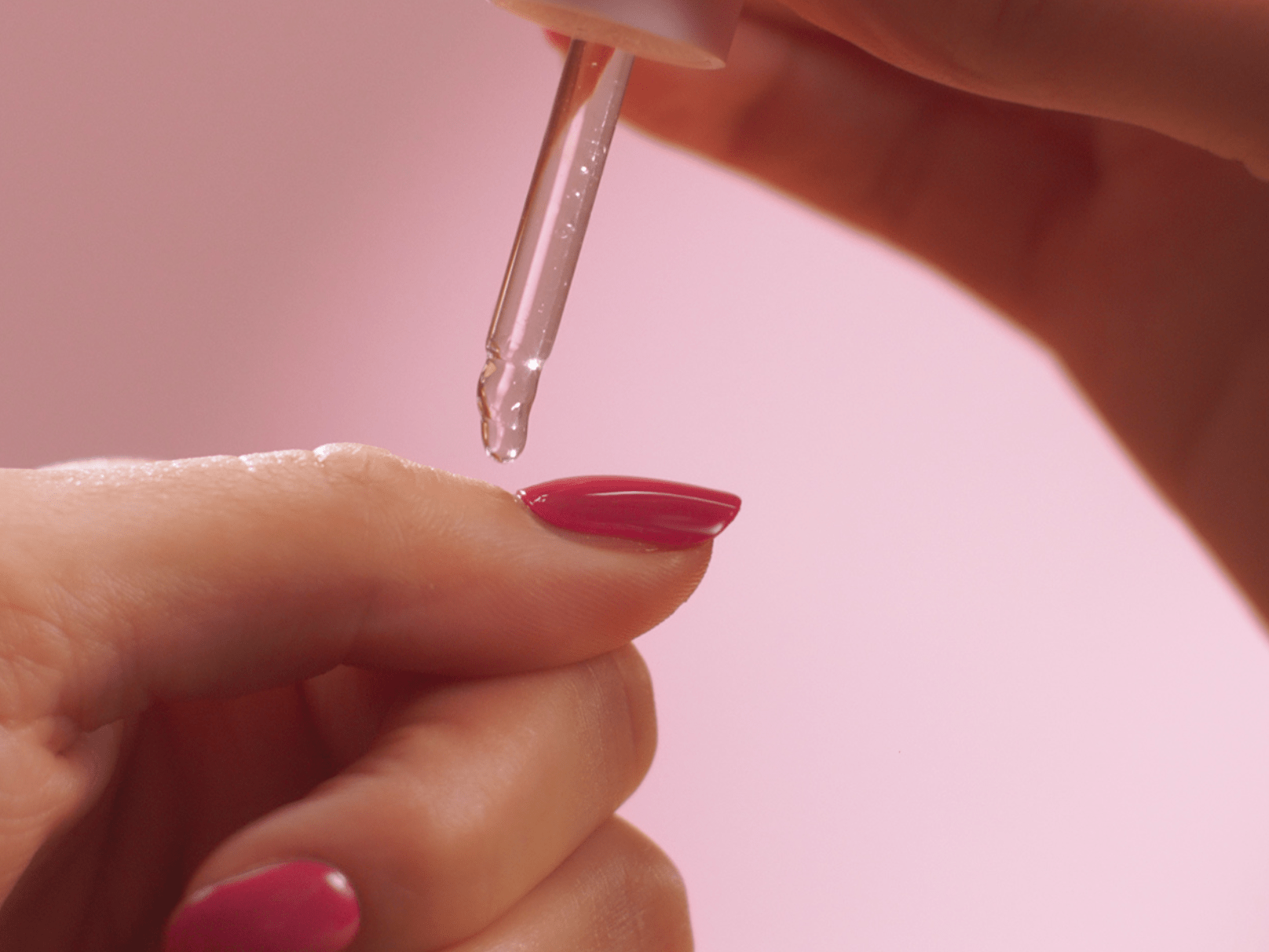 Hand with pink nail polish, dripping skin care oil on fingers with a pipette against a pink background.